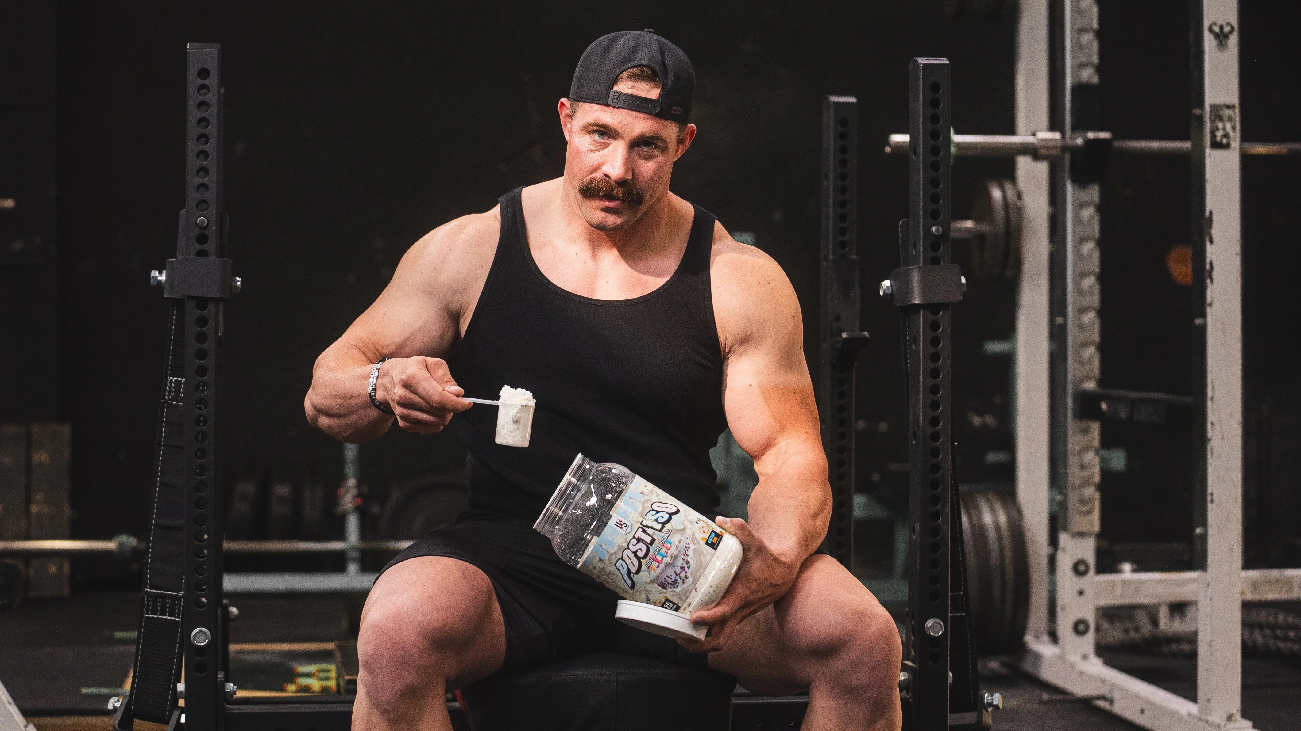Lifter scooping pre-workout from a tub while seated in a squat rack