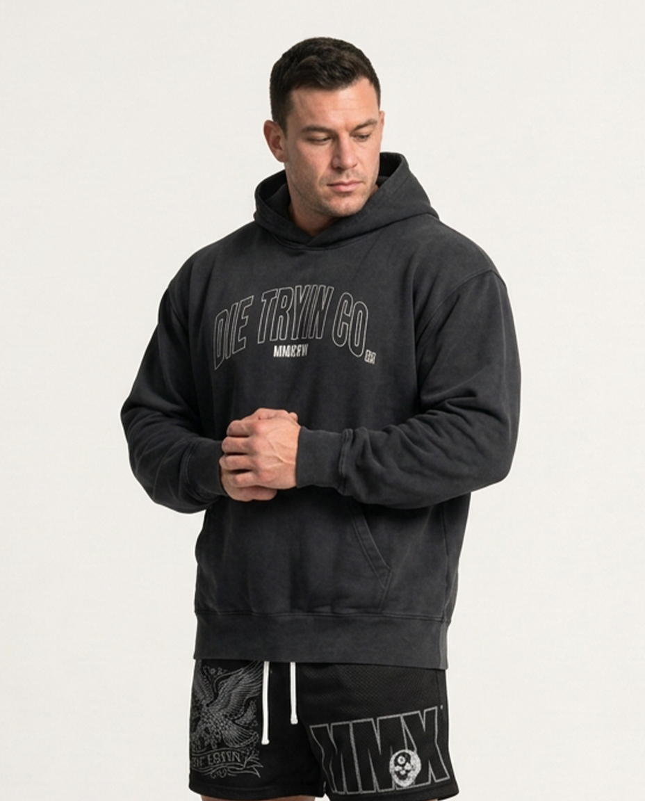 Man in a washed black hoodie with an outlined, arched DIE TRYIN CO. chest print, worn with black graphic mesh shorts