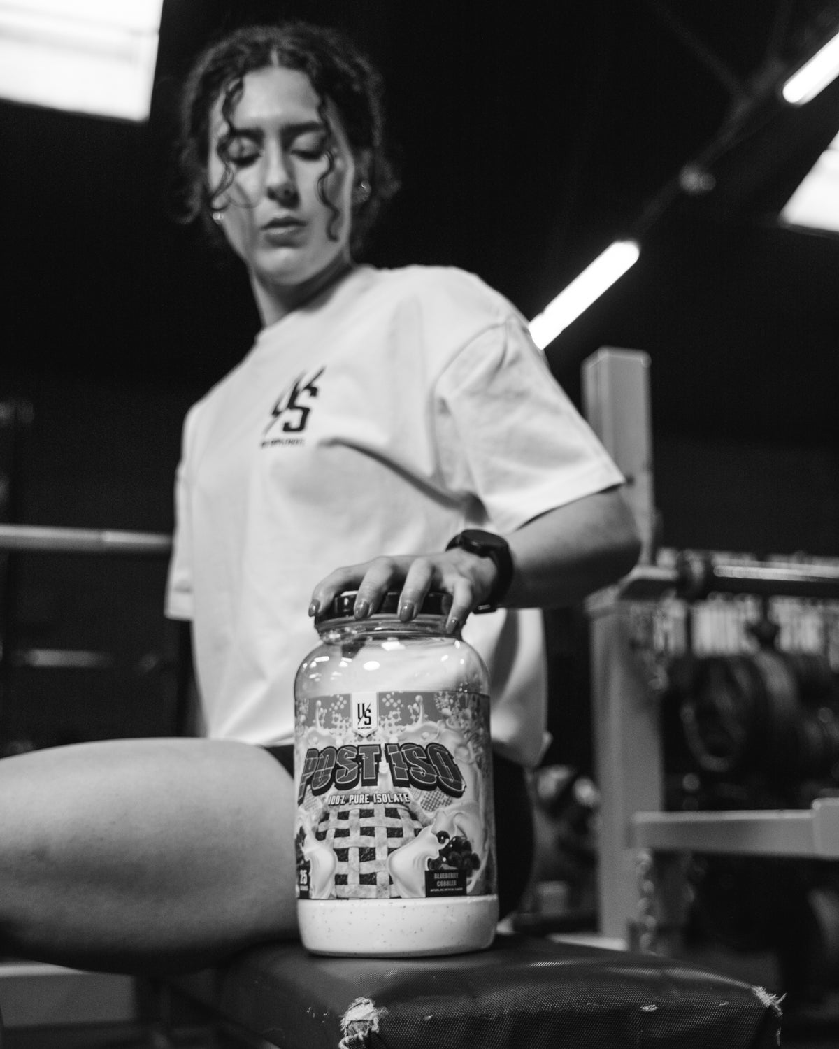 Black-and-white photo of a woman in a white tee on a gym bench with her hand on a Post Iso protein tub