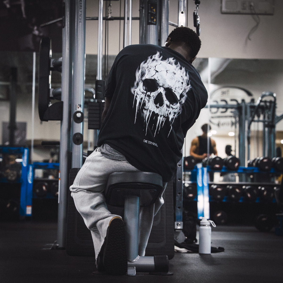Lifter in grey sweatpants and a black tee with a flaming skull back print, kneeling at a cable machine in a gym