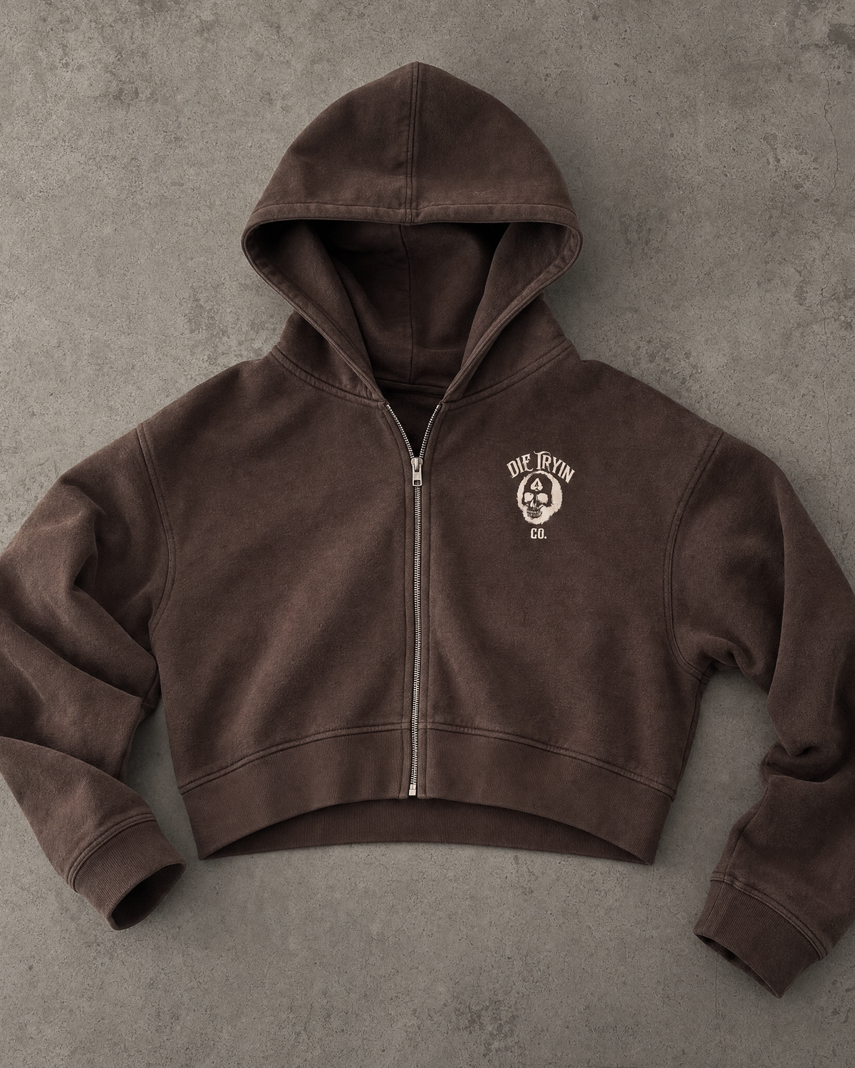 Brown cropped zip-up hoodie with a small skull Die Tryin Co. chest logo, flat lay on concrete