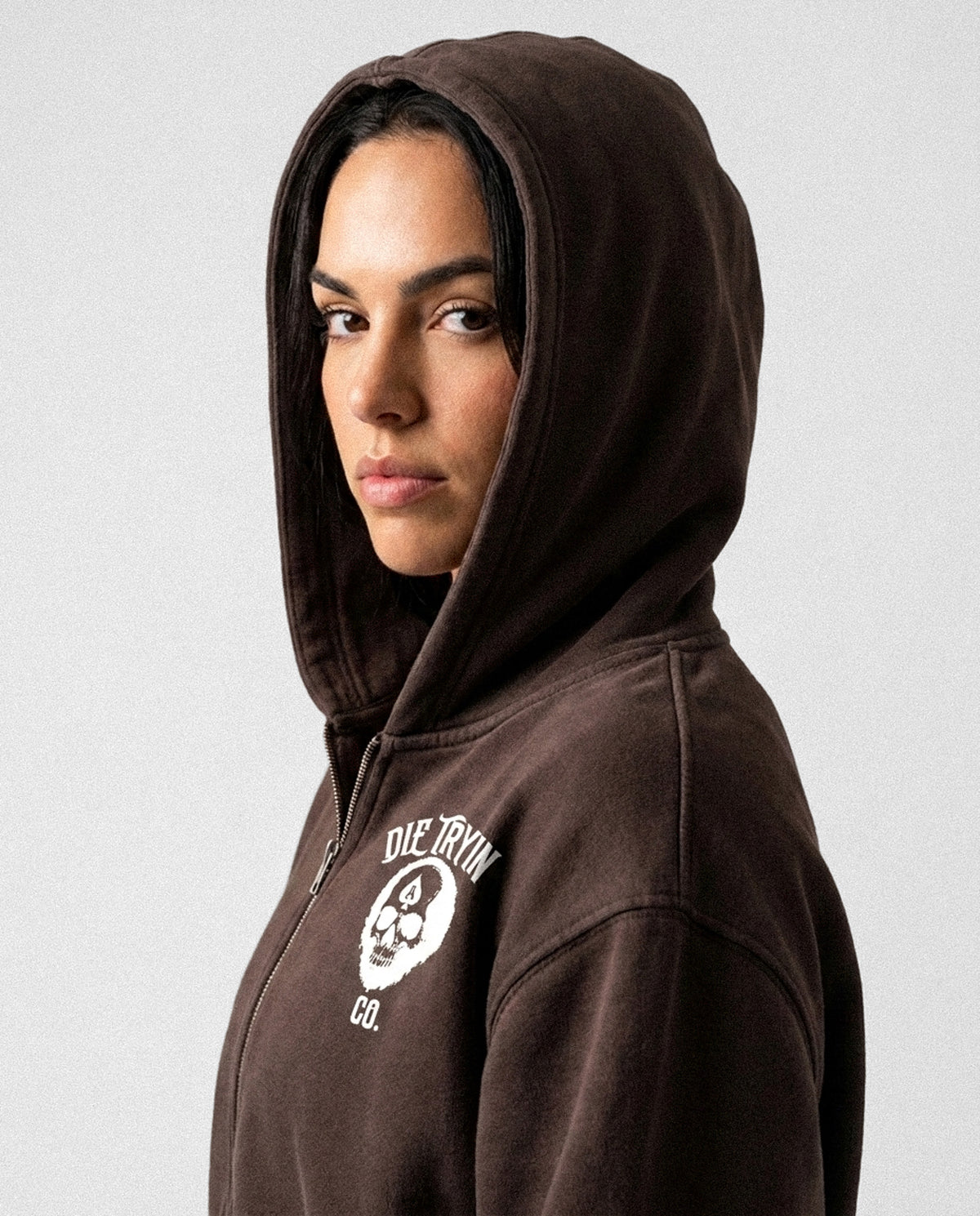 Close-up of a woman in the hood of a brown zip-up hoodie, cream Die Tryin Co. skull logo printed on the chest