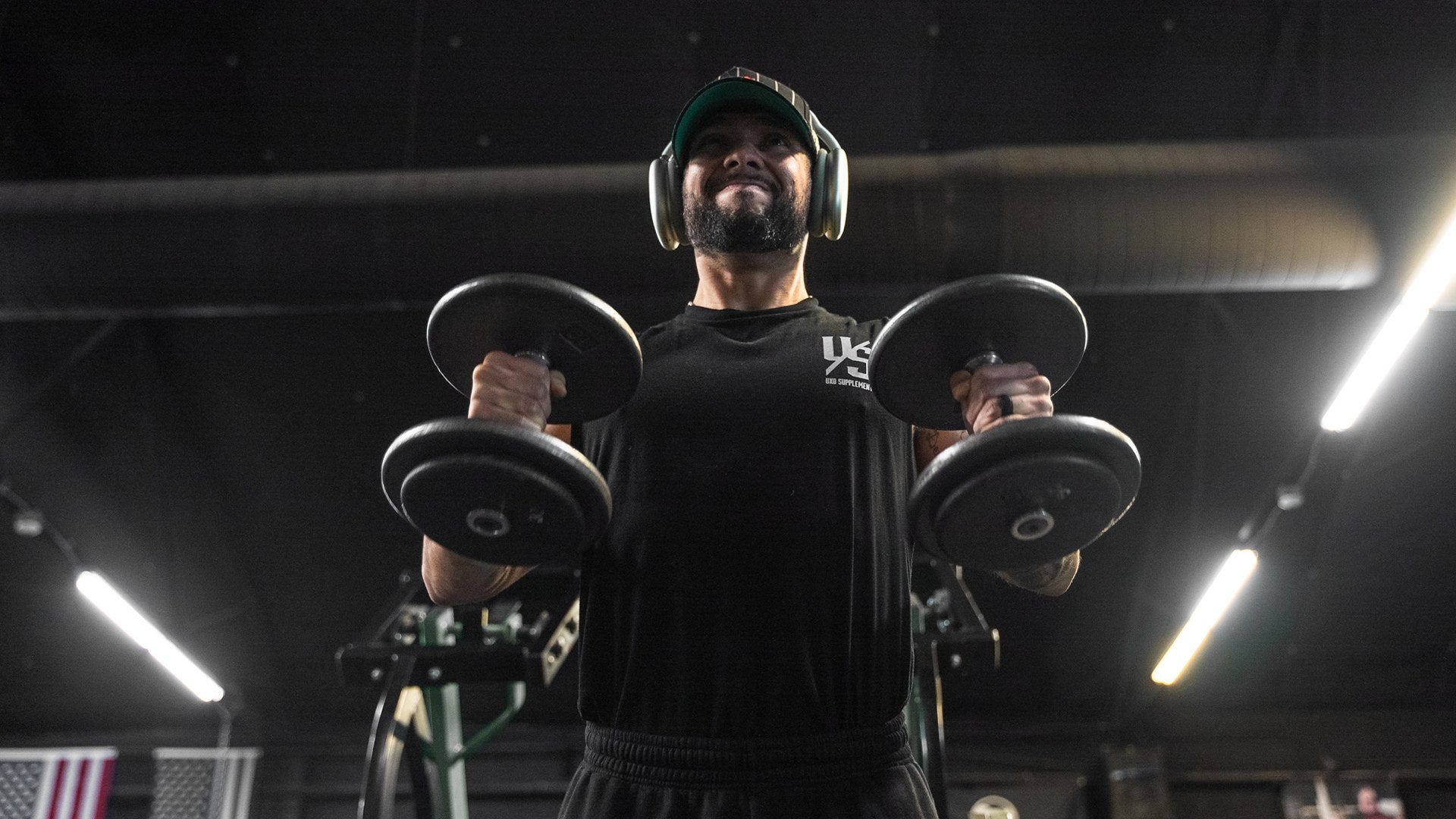 Lifter in headphones holding two dumbbells under gym lights