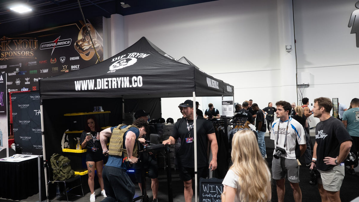 Die Tryin Co. booth at the 2026 Olympia expo: a lifter in a weighted vest on the dip bar under the WWW.DIETRYIN.CO canopy