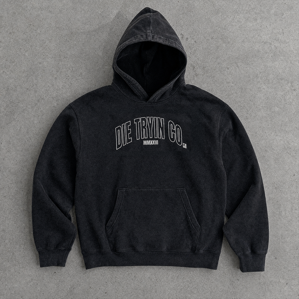 Black hoodie with an arched outlined Die Tryin Co. MMXXVI print, flat lay on concrete