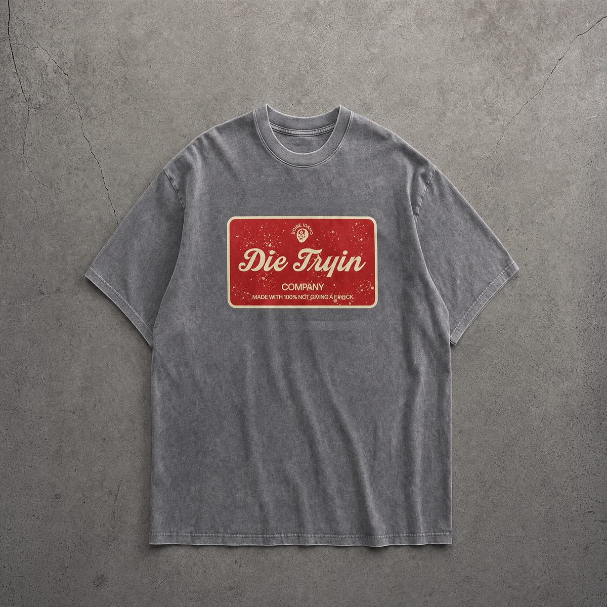 Washed grey oversized tee with a red Die Tryin Company label graphic, flat lay on concrete