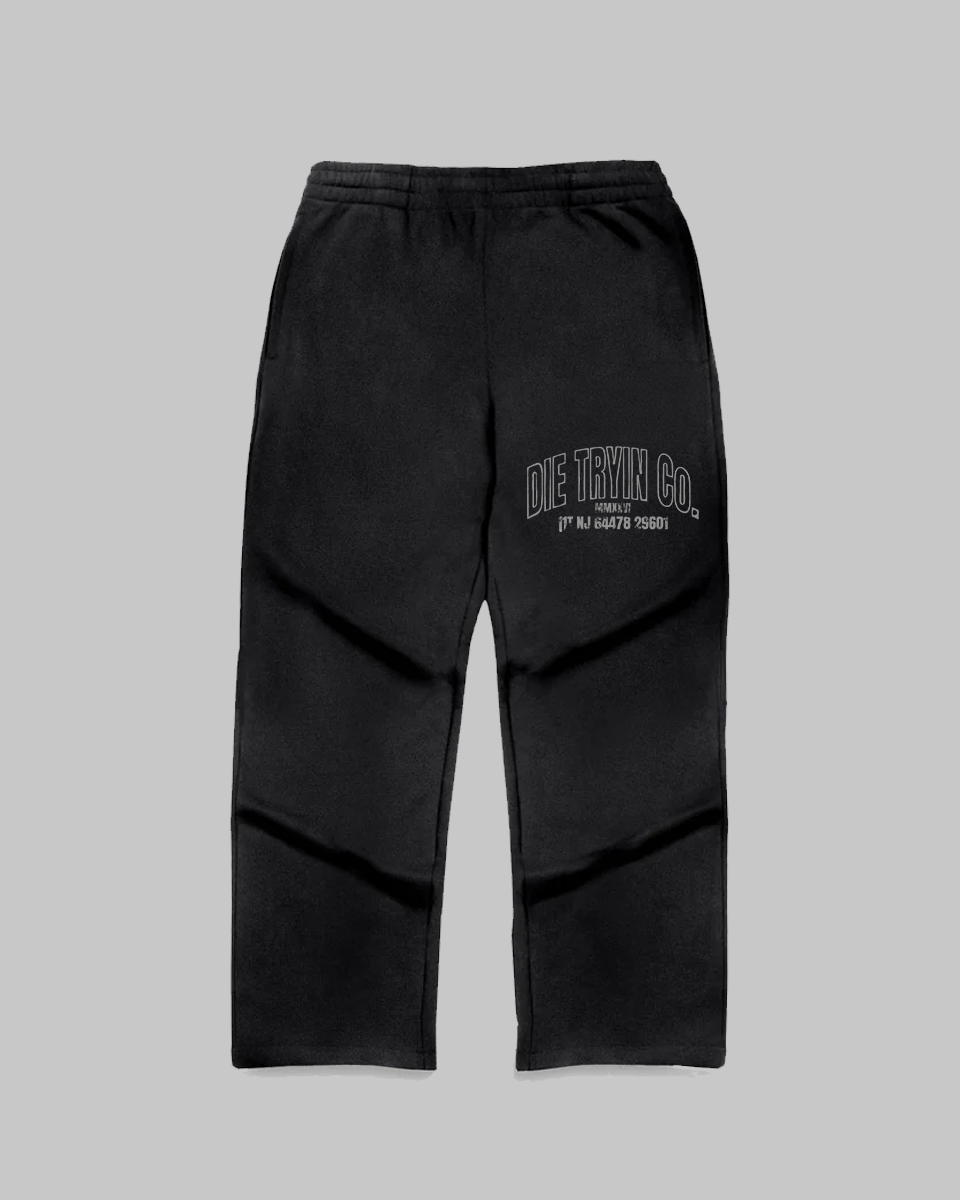 Black straight-leg joggers with an arched Die Tryin Co. print on the left thigh, front view on grey