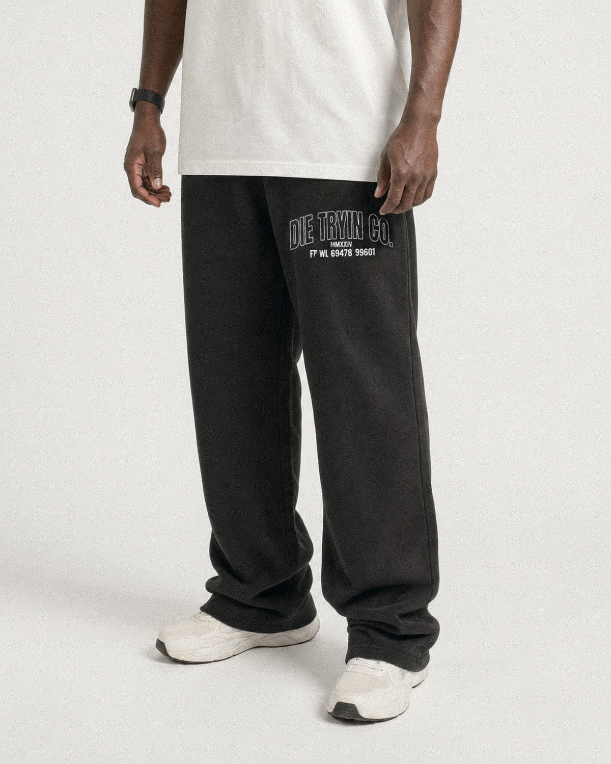Man in a white tee and sneakers wearing black straight-leg joggers with an arched Die Tryin Co. thigh print
